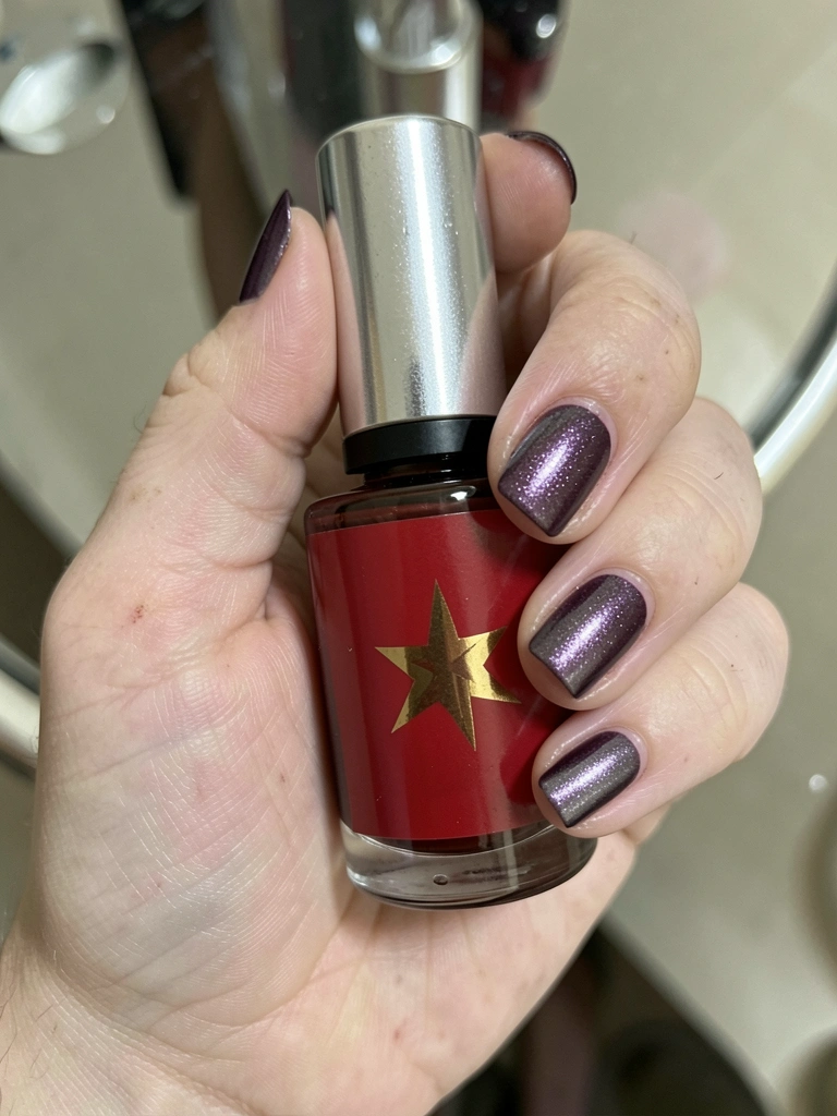 Mãos de pele clara com unhas pintadas em roxo metálico e cinza chumbo sobre fundo neutro.